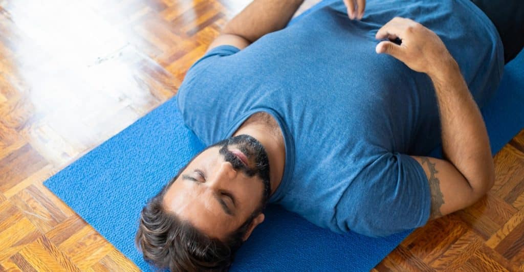 A man is lying on a blue yoga mat with his eyes closed and hands resting on his stomach, appearing to be meditating or practicing deep breathing.