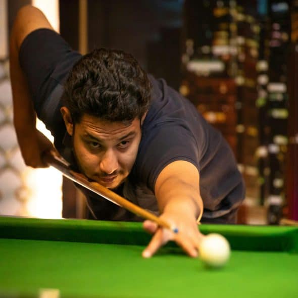 A man with dark hair and a focused expression aims a pool cue at a white ball on a green pool table.