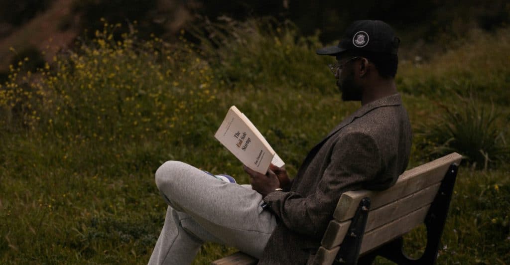 A man wearing a blazer, t-shirt, and a baseball cap is sitting on a park bench reading a book, with blurred greenery in the background