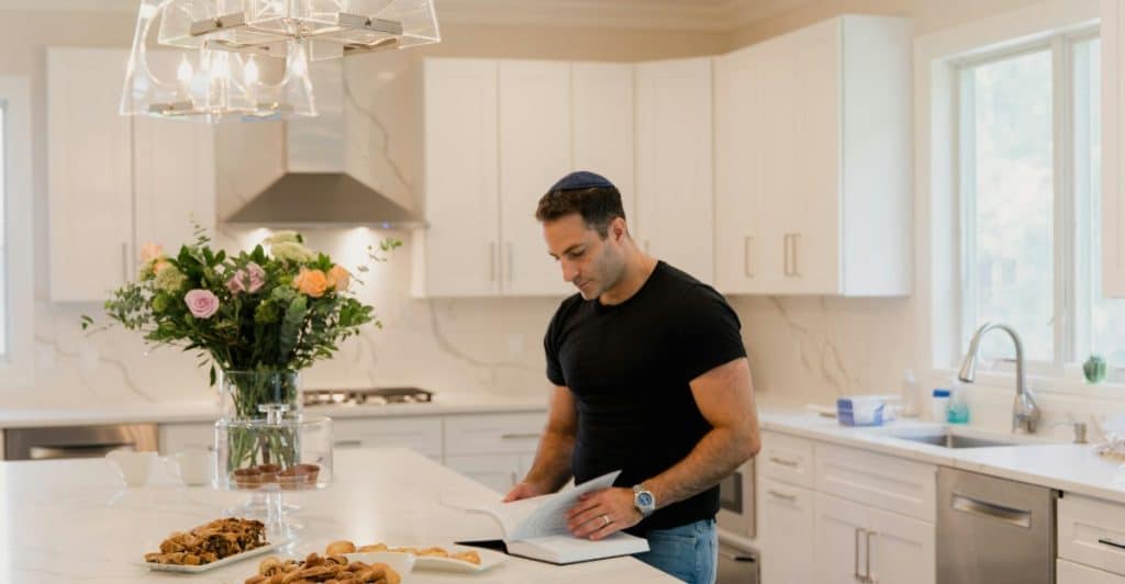 A man is standing in a kitchen, reading a book, with baked goods and a flower pot on the counter.