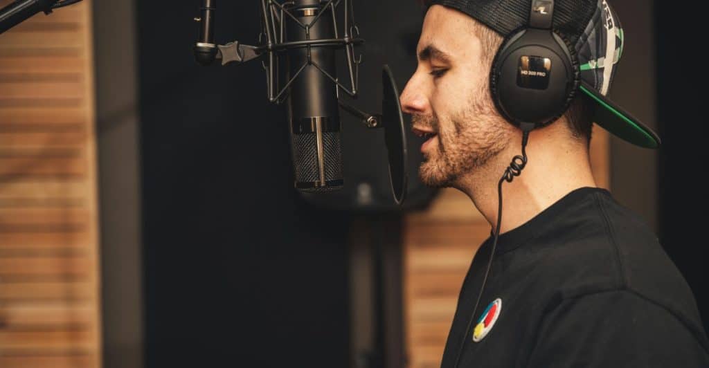 A man wearing headphones and a baseball cap speaking into a studio microphone in a recording booth.