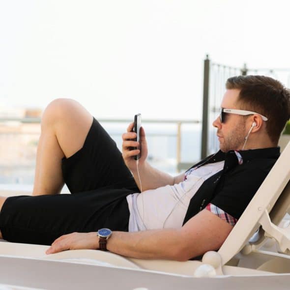 A man wearing sunglasses and headphones, relaxing on a lounge chair by a pool or balcony and looking at his phone