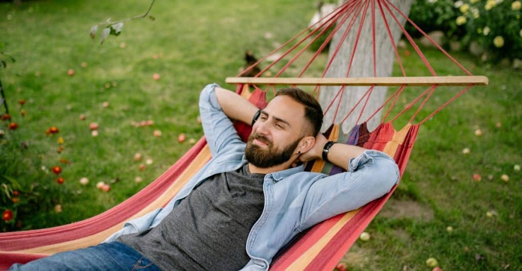 A man with a beard lies relaxing in a red and yellow hammock outdoors, with trees and fruit visible around him.