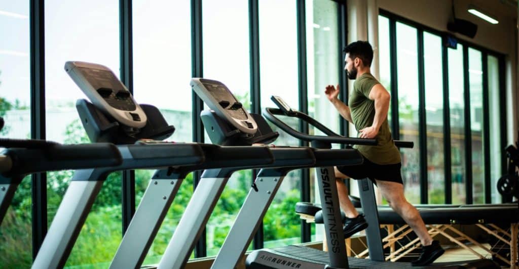 Man running on a treadmill in a modern gym with large windows overlooking greenery.