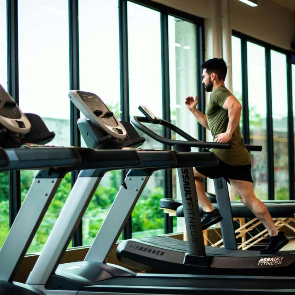 Man running on a treadmill in a modern gym with large windows overlooking greenery.