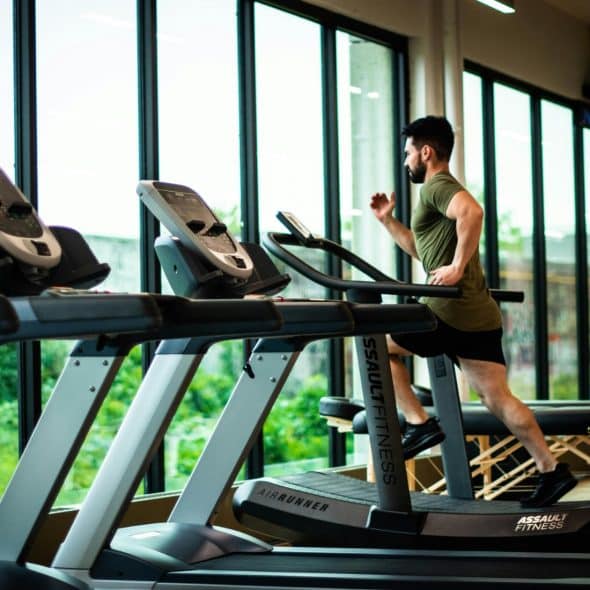 Man running on a treadmill in a modern gym with large windows overlooking greenery.