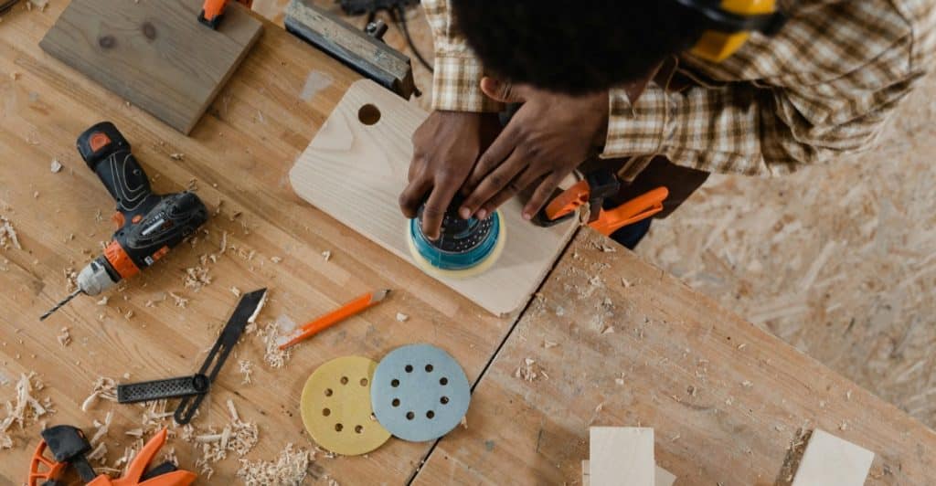 Aerial view of a person sanding a wooden board with an orbital sander on a workbench, surrounded by wood shavings and other tools.