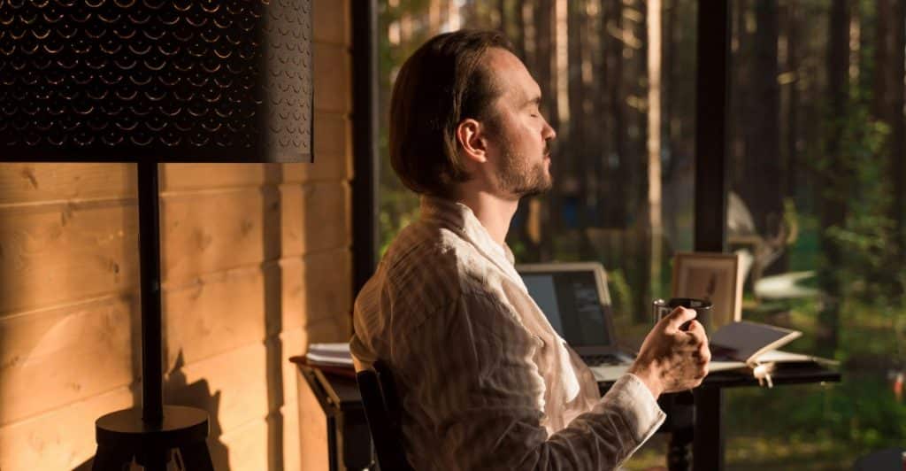 A man sitting at a table in a room with large windows, holding a mug and looking contemplative.