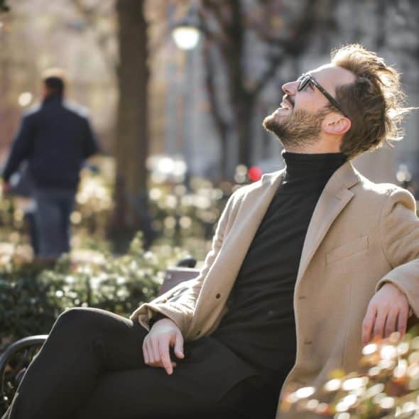 A man in a tan coat and black turtleneck, wearing glasses, sits on a park bench bathed in sunlight, looking up and smiling.