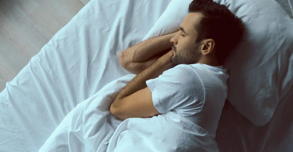 An overhead view of a man sleeping on his side in a bed with white sheets and pillows.
