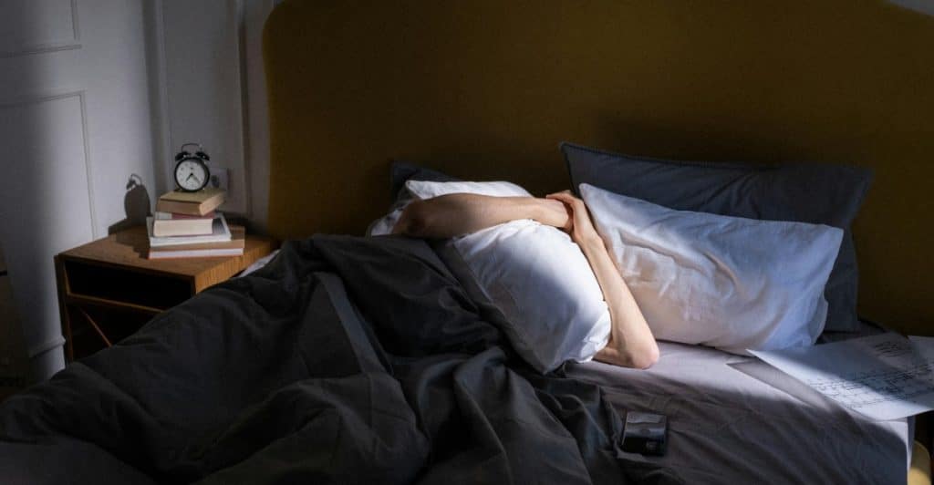 A person is lying in bed covering their head with a pillow, with an alarm clock and books on a nightstand beside the bed.
