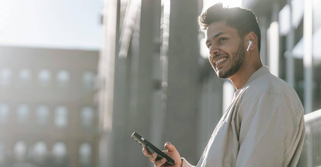 A man is smiling and holding a smartphone, with earbuds in his ears, in an urban setting.