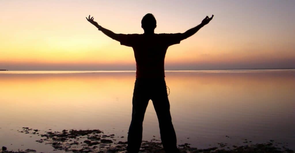 A silhouette of a person standing by a body of water with arms raised towards the sunset.