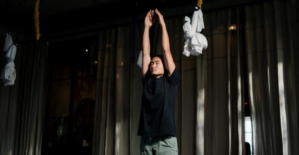 A man standing with his arms raised above his head, palms together, in what appears to be a studio or gym setting.