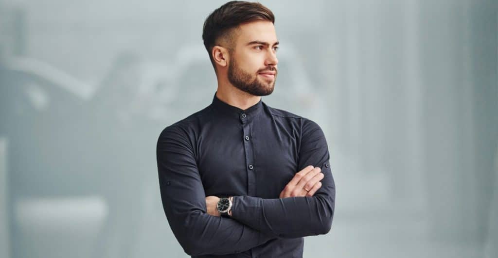 A man with a neatly trimmed beard and dark, slicked-back hair stands with his arms crossed, wearing a dark button-up shirt and a watch, looking off to the side.