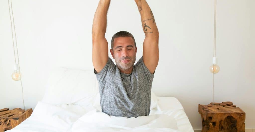 A man with a tattoo on his arm is sitting up in bed, stretching his arms overhead with his eyes closed.
