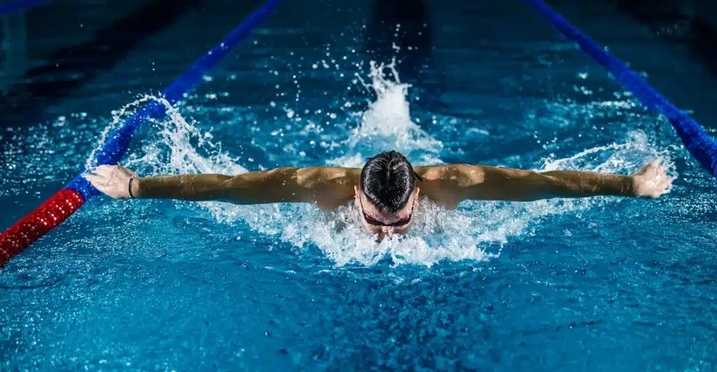A muscular man with dark hair and goggles swimming the butterfly stroke in a blue swimming pool with lane markers.