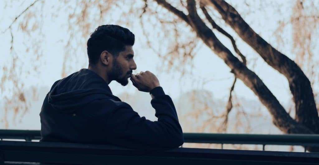A man sitting pensively on a bench outdoors with a bare tree in the background.