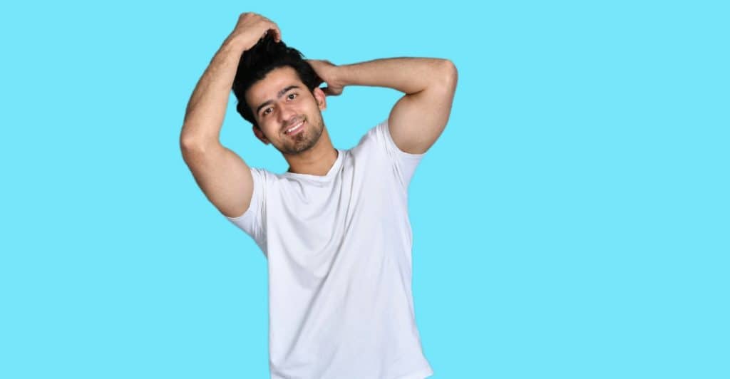A man in a white t-shirt is touching his hair with both hands against a blue background.