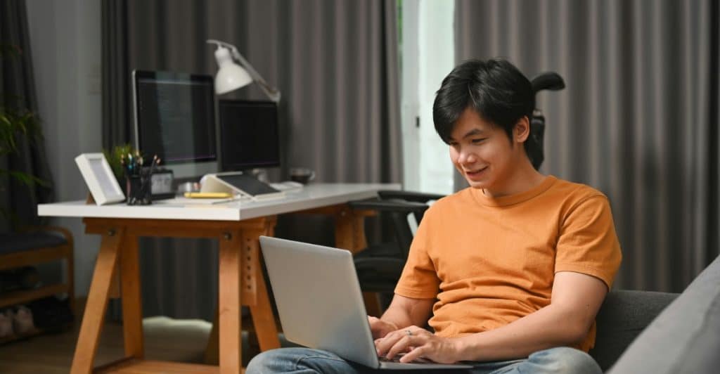 A man sitting comfortably on a couch with a laptop, smiling and appearing to be working or learning, with a desk setup in the background.