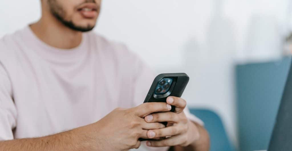A person holding and looking at a smartphone while sitting at a table with a laptop and papers, relating to managing digital information like passwords.