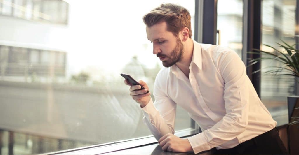 A man in a white shirt leans against a window using a smartphone.