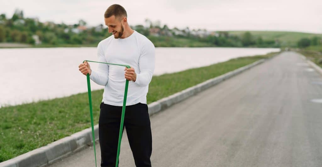 A muscular man in a white shirt using a green resistance band on a path.