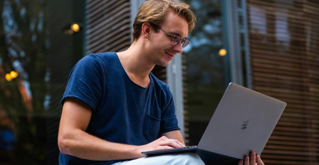 A man with glasses is shown using a laptop while sitting outside in a casual setting.