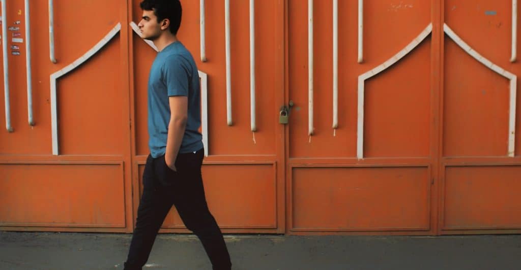 A man in a blue t-shirt and black pants walking  down a street in front of an orange building with patterned walls and a locked gate.