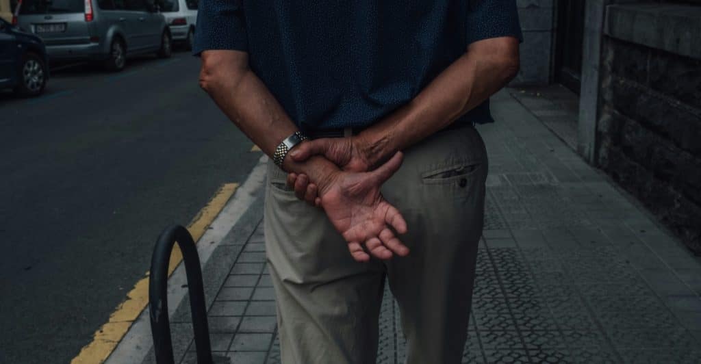 A view from behind a man walking away on a sidewalk next to a street with parked cars. He is wearing a dark polo shirt and tan pants and has his hands clasped behind his back.