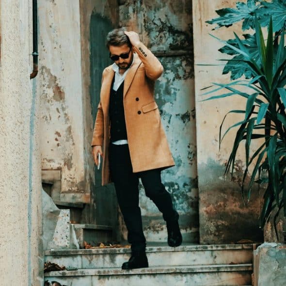 A stylish man in a tan coat walks down outdoor steps with his hand on his head.
