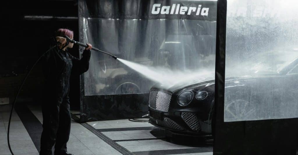 A person wearing headphones uses a pressure washer to clean a black car inside a car wash stall.