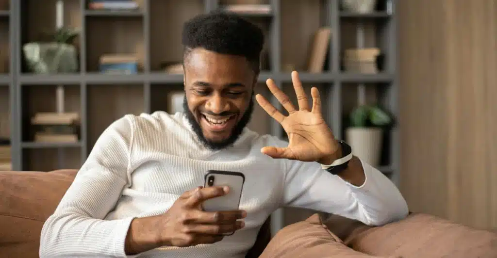 A man is smiling and waving while looking at his smartphone, sitting on a couch indoors.