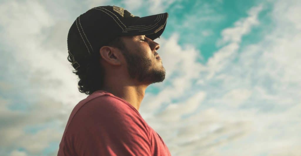 Side profile view of a man wearing a baseball cap, looking up at the sky with his eyes closed or partially closed.