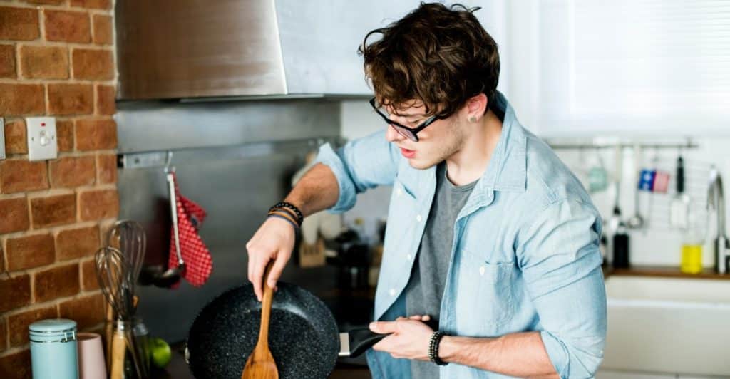 A man wearing glasses and a light blue button-up shirt over a grey t-shirt is cooking in a kitchen, using a wooden spatula to transfer something from a black frying pan to a white plate.