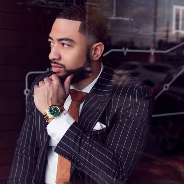 A man wearing a pinstripe suit and tie with a gold-colored watch, hand on his chin, looking away from the camera sitting behind a window.