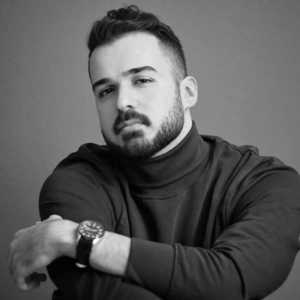 Black and white photo of a man with a beard, wearing a turtleneck and watch, sitting with arms crossed.