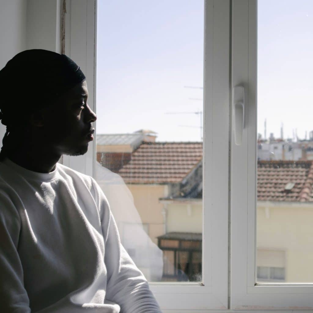 A young Black man in a white sweatshirt gazes pensively out a window overlooking a cityscape.