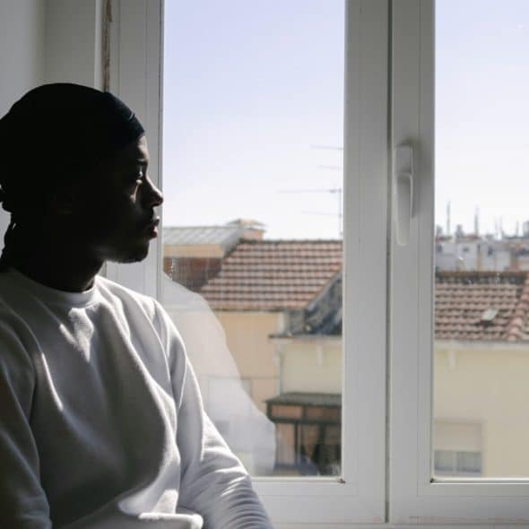 A young Black man in a white sweatshirt gazes pensively out a window overlooking a cityscape.
