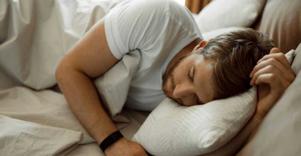 A man with a beard is sleeping on his side in a bed, covered by a white duvet.
