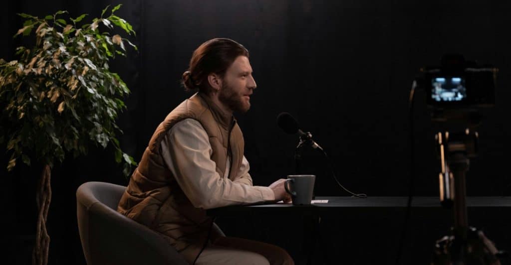 A man with a beard and hair tied back sitting at a table with a microphone and a mug, wearing a light-colored shirt and a puffer vest with a camera on a tripod in front of him and a plant to his left.