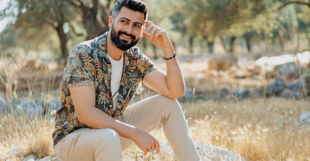 Man with a beard sitting on rocks outdoors, wearing a patterned short-sleeved shirt, khaki pants, and white sneakers and smiling at the camera with one hand near his temple.