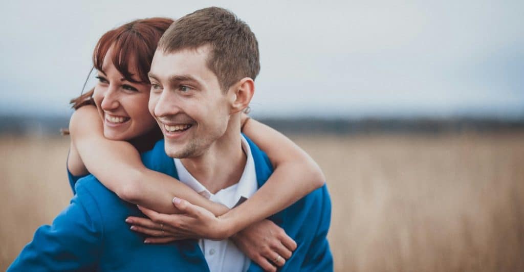 A cheerful couple embraces in a field, with one person giving the other a piggyback ride.