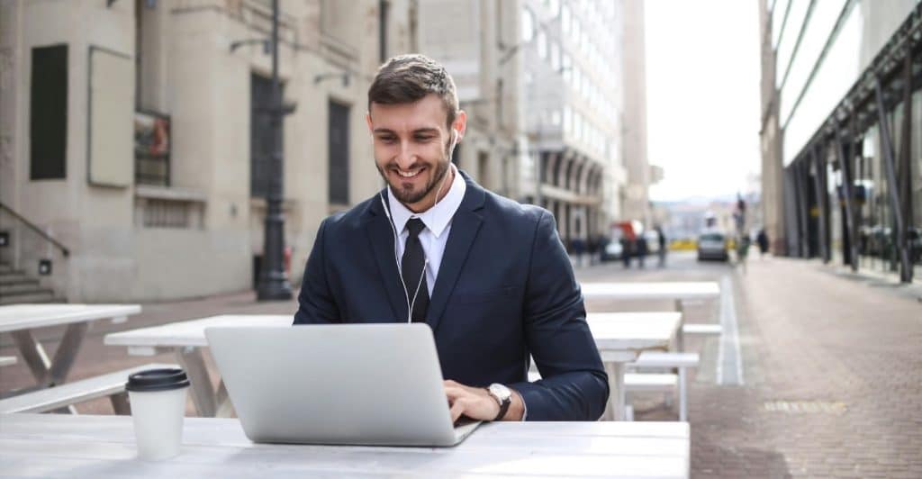 A smiling man in a suit working on a laptop wearing earphones at an outdoor cafe table.