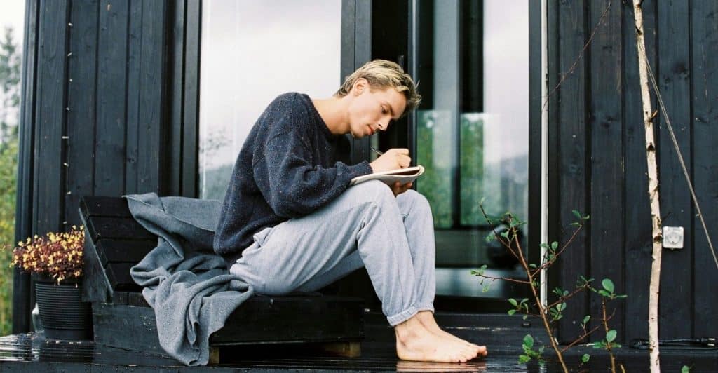 A man sitting on a porch writing in a notebook in front of a modern cabin.