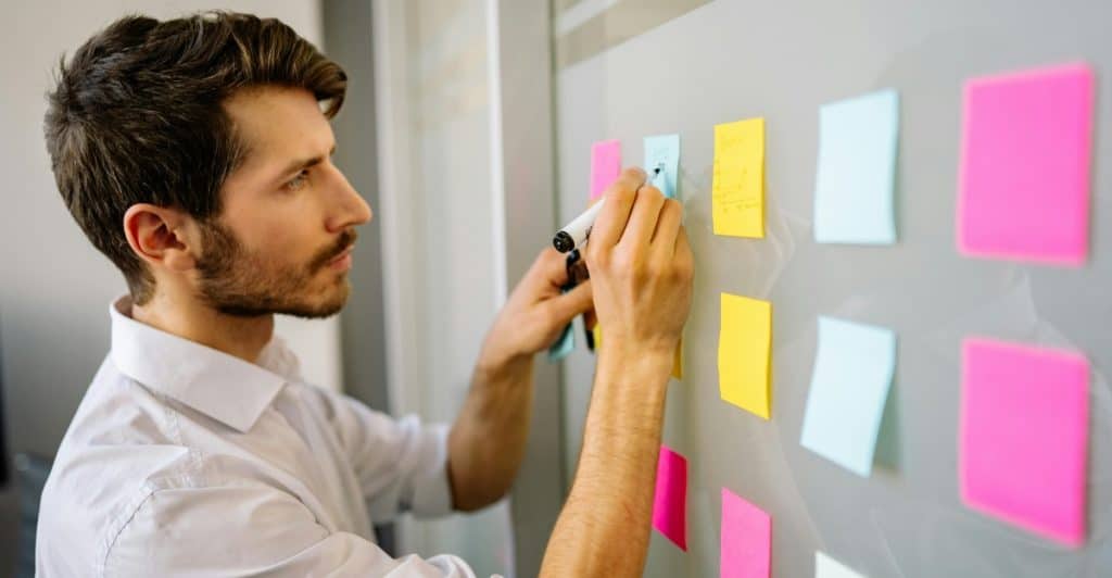 A man in a white shirt is writing on colorful sticky notes placed on a glass wall or window.