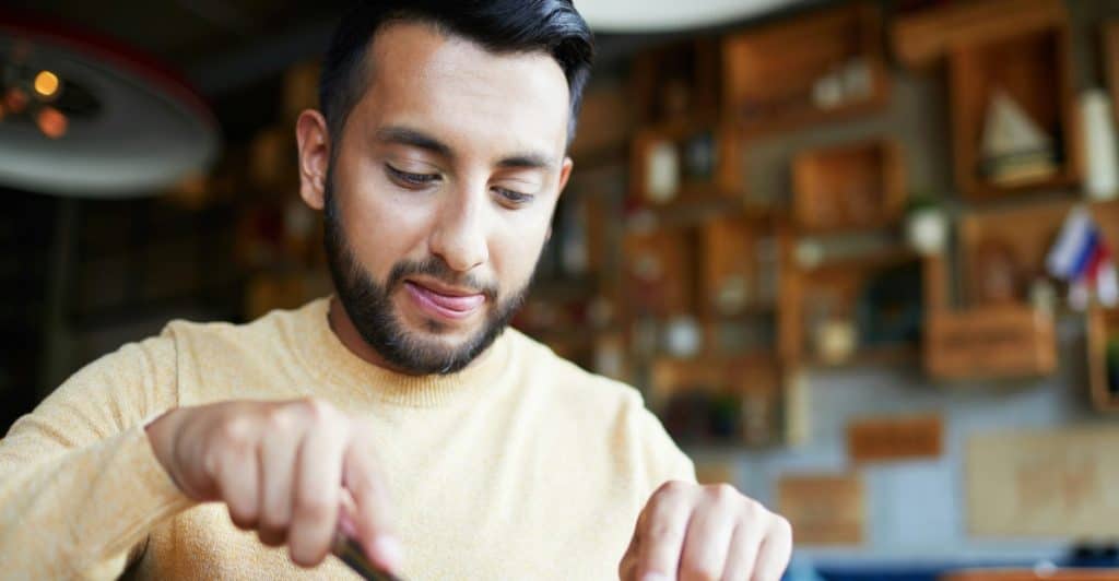 A man with dark hair and a light yellow sweater is holding a knife and fork, preparing to eat a pizza.
