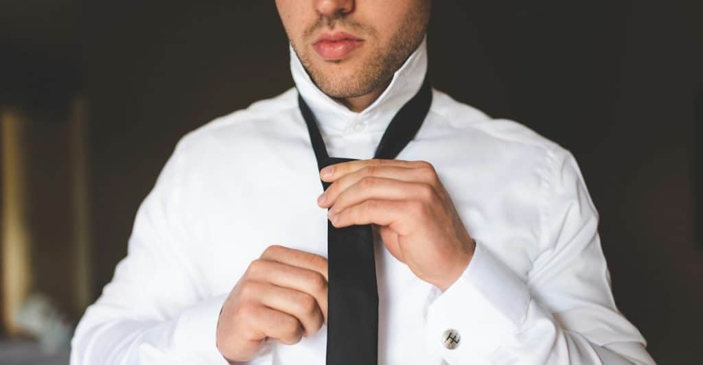 A close-up of a man wearing a white collared shirt and adjusting a black tie.