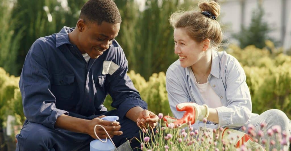 A man and a woman are kneeling side-by-side in a garden, wearing gloves and tending to plants, looking at each other and smiling.