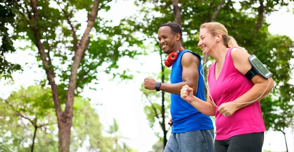 A man in a sleeveless shirt and a woman in a pink top jogging together with trees in the background.